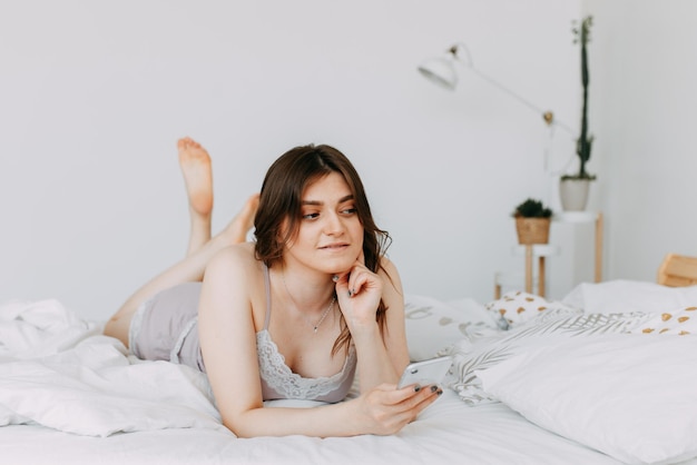 Фото Счастливая девушка в пижаме делает селфи, общается с помощью телефона и технологий в постели дома.