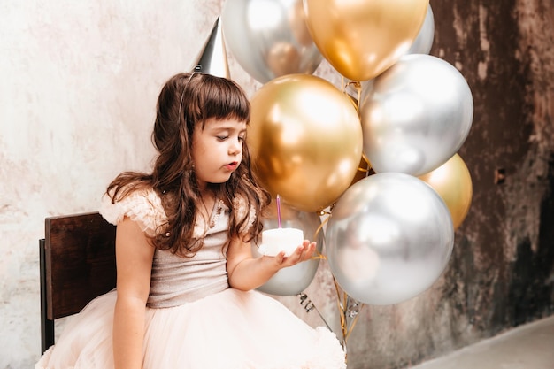 Счастливая маленькая девочка загадывает желание и задувает свечи на воздушных шарах для торта. место для текста, декоративная стена, день рождения.