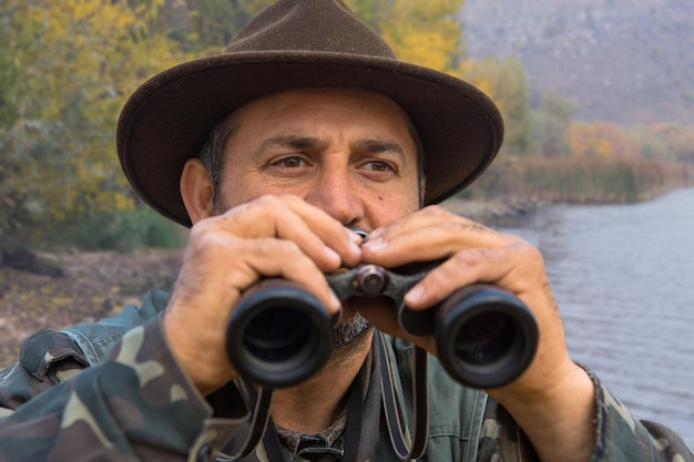 사진 쌍안경을 쓴 모자를 쓴 사냥꾼이 숲을 배경으로 먹이를 찾습니다