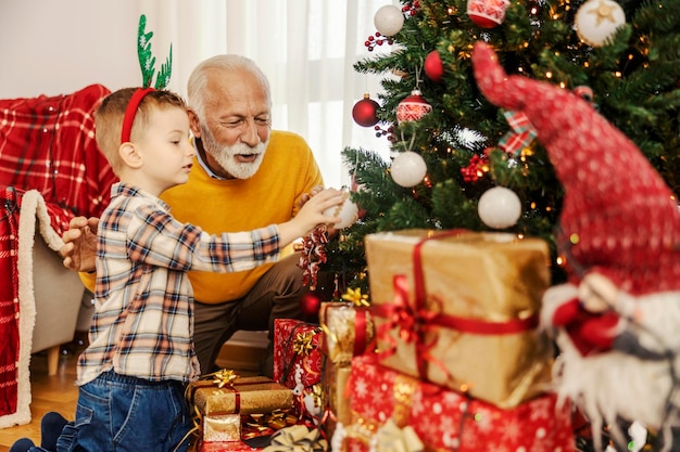 Фото Веселый дедушка играет с рождественскими украшениями со своим внуком возле рождественской елки