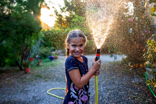 写真 落ちる太陽の金色の光の下で庭のホースで水を噴射する喜びに満ちた女の子が遊んでいます