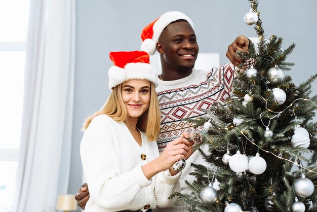 즐거운 다민족 커플이 함께 크리스마스 트리를 장식하고 카메라를 바라보고 있습니다. 백인 여자와 산타 모자를 쓴 아프리카계 미국인 남자가 함께 크리스마스를 축하합니다.