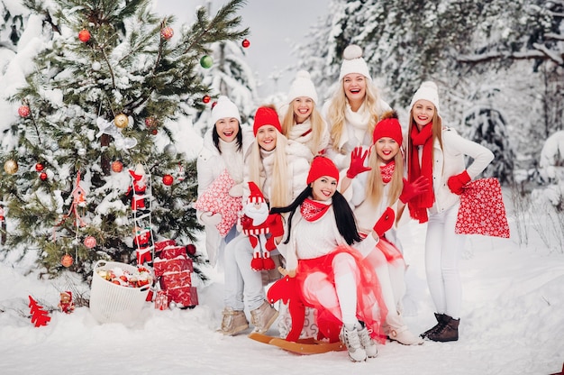 Большая группа девушек с рождественскими подарками в руках стоит в зимнем лесу. девушки в красно-белой одежде с рождественскими подарками в заснеженном лесу.