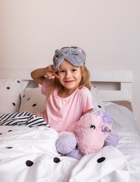 Маленькая блондинка в пижаме и с маской для сна сидит на кровати с мягкой игрушкой