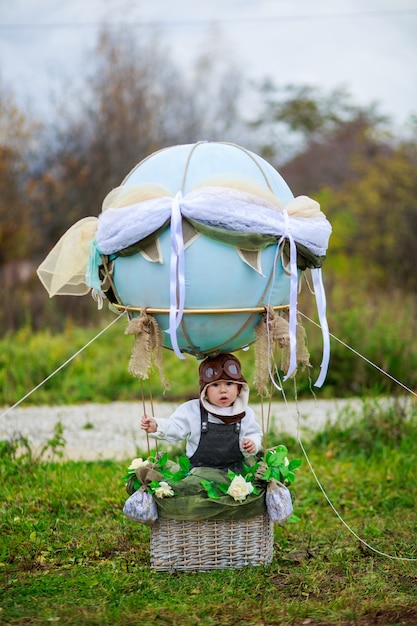 写真 飛行士の帽子をかぶった小さな男の子は、フィールドのおもちゃの風船のバスケットに座っています。