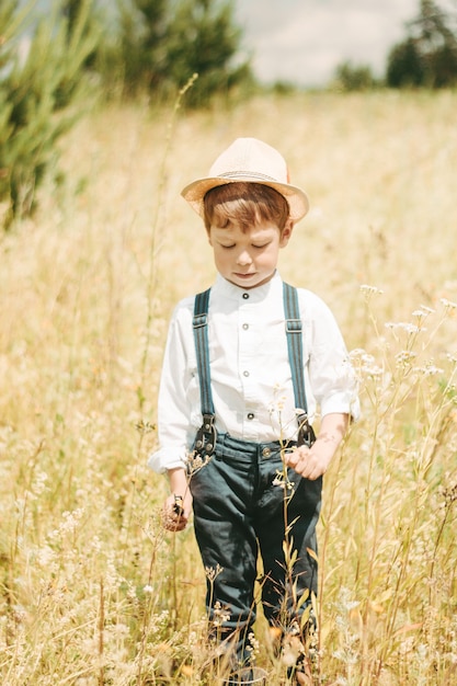 写真 小さな農夫が夏の畑を歩いていて、小さな男の子が麦わら帽子をかぶっています。ゴム長靴と白いシャツを着た少年。カントリースタイル