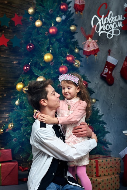 写真 少女と少年が大晦日とクリスマスのクリスマスツリーの近くを抱き締めます。背景のロシア文字の言葉：明けましておめでとう。