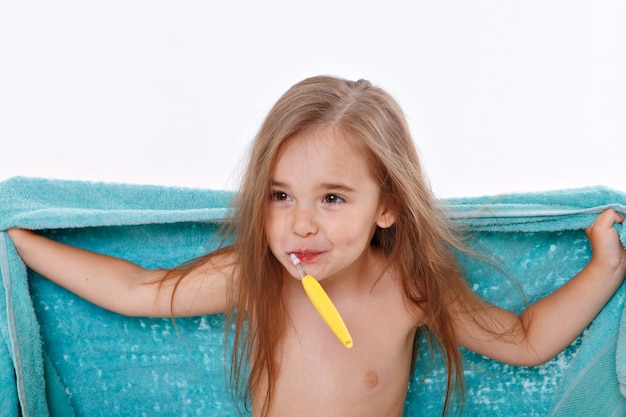 Фото Маленькая девочка чистит зубы на белом фоне. портрет ребенка с желтой зубной щеткой. синее полотенце на шее. утренние гигиенические процедуры