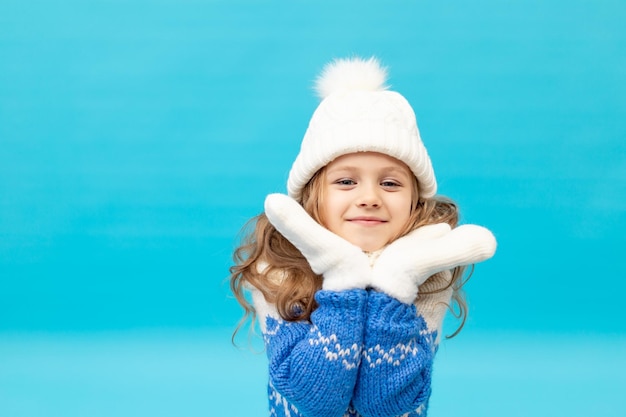 Фото Маленькая девочка на синем фоне в зимней шапке и свитере с варежками улыбается место для текста