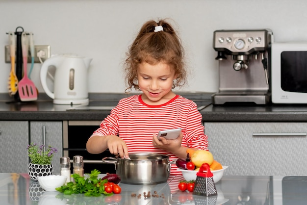 小さな女の子が携帯電話を持って、新鮮な野菜から何を調理するかを考えます。