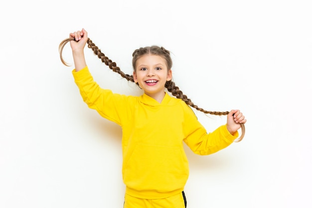 Фото Маленькая девочка держит свои длинные волосы в косах в руках и широко улыбается ребенок в желтом спортивном костюме на белом изолированном фоне