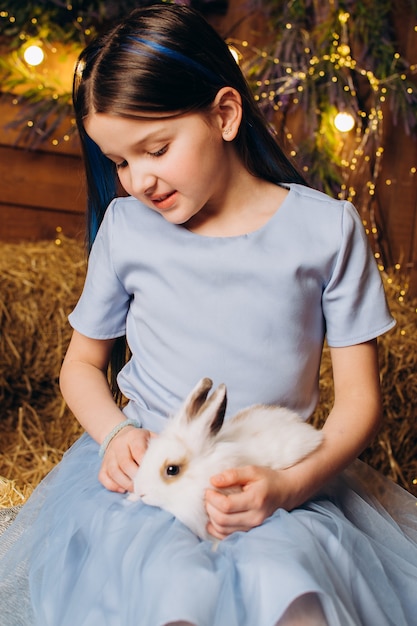 사진 파란 드레스를 입은 어린 소녀가 토끼와 함께 농장에 앉아 있다