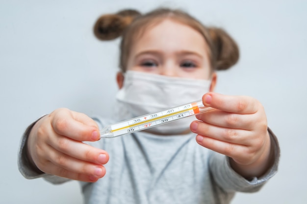 医療マスクの少女は、温度計を示しています。疫学期間、呼吸器疾患および高温の医療概念。コロナウイルスの流行防止