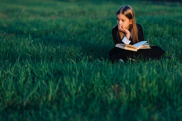 Маленькая девочка в черном сидит на траве и держит зеленую книгу в свете заходящего солнца. ребенок задумчиво смотрит вдаль на природе на природе.