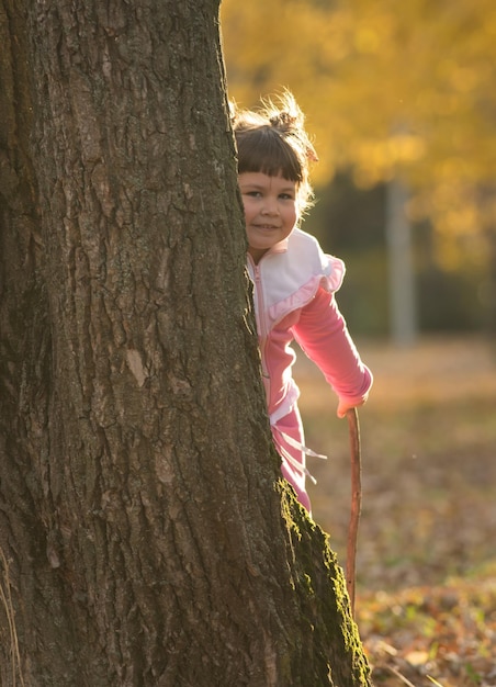 Фото Маленькая девочка в розовом костюме прячется за деревом в осеннем парке