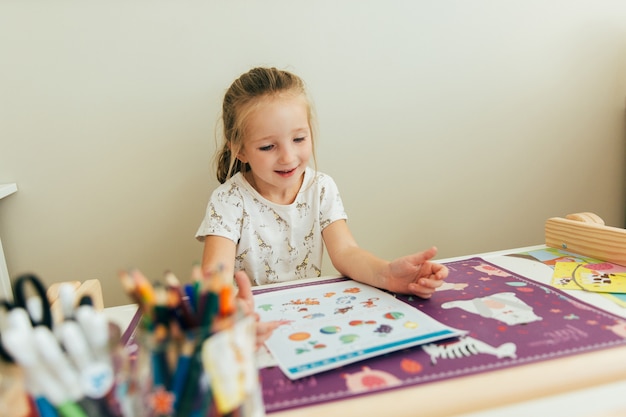 写真 小さな女の子は彼の机に座って学ぶことを幸せです。ホームスクールのコンセプト。教育コンセプト。子供の学習の背景。幼児の手作りゲーム。幼稚園教育。