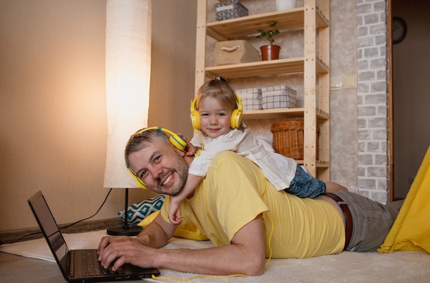 Фото Маленькая девочка лежит на спине отца и слушает музыку в желтых наушниках, глядя на свой ноутбук. концепция семейного счастья