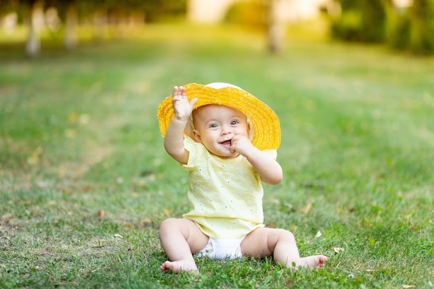 사진 8 개월의 어린 소녀는 노란색 여름 드레스와 모자에 푸른 잔디에 여름에 앉아있다