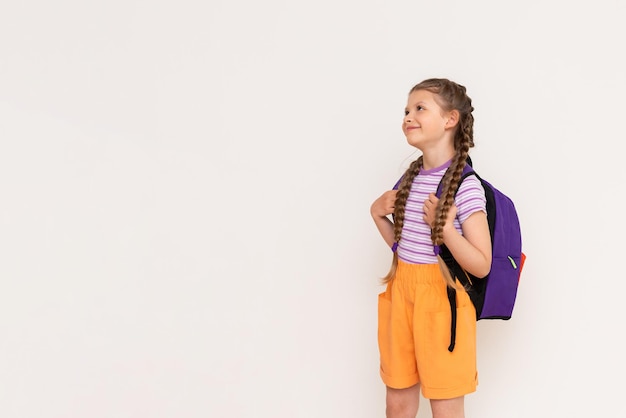 가방을 등에 메고 있는 어린 소녀가 흰색으로 격리된 배경에서 귀하의 광고를 보고 있습니다. 어린이를 위한 여름 코스 복사 공간