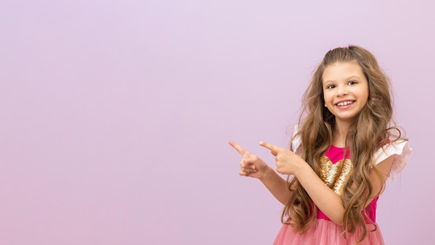 Фото Маленькая девочка с вьющимися волосами в красивом розовом платье показывает пальцем на вашу рекламу.