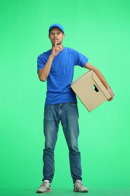 Фото Мужчина-доставщик на зеленом фоне в полной высоте с коробкой показывает знак молчания