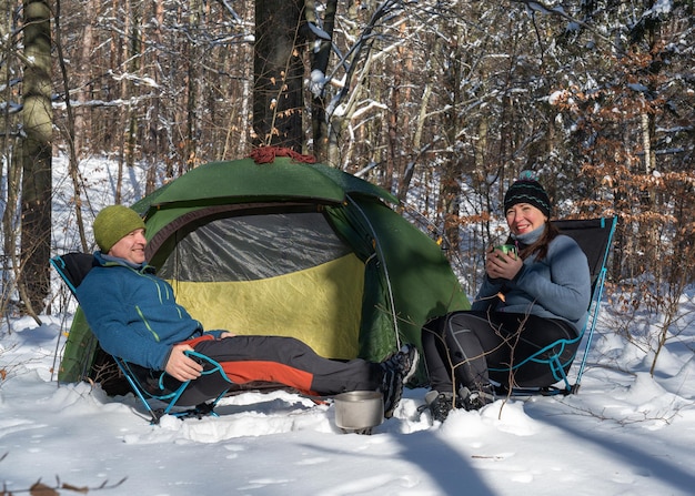 写真 テントの近くの雪に覆われた森の椅子に座っている男性と女性 お茶の準備 活動的な生活 人生の喜びと満足 冷ややかな空気が燃える食べ物 晴れた日 たくさんの雪