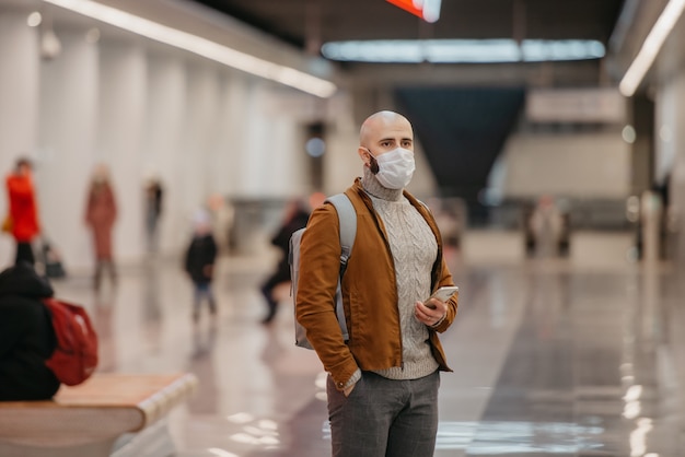 Фото Мужчина в медицинской маске держит смартфон и смотрит в сторону, ожидая поезда в центре станции метро. лысый парень в хирургической маске сохраняет социальную дистанцию.