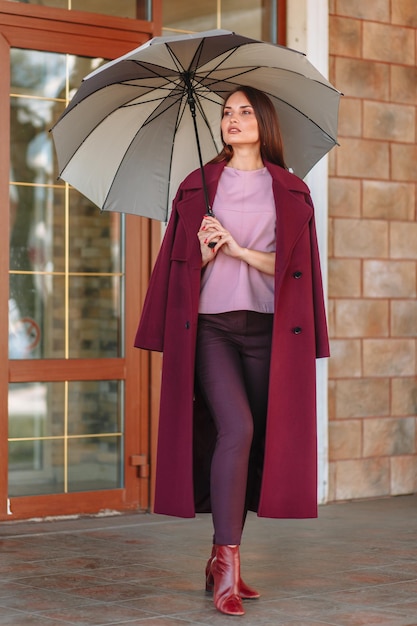 Девушка-модель демонстрирует модную верхнюю одежду с зонтиком в руке на улице на веранде летнего кафе одежда для выставочного зала