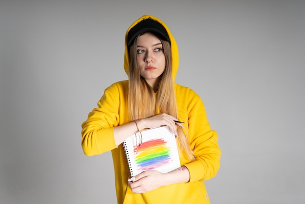 写真 黄色いパーカーと黒い帽子をかぶった物思いにふける女性は、lgbtqの虹が描かれたノートを持っています
