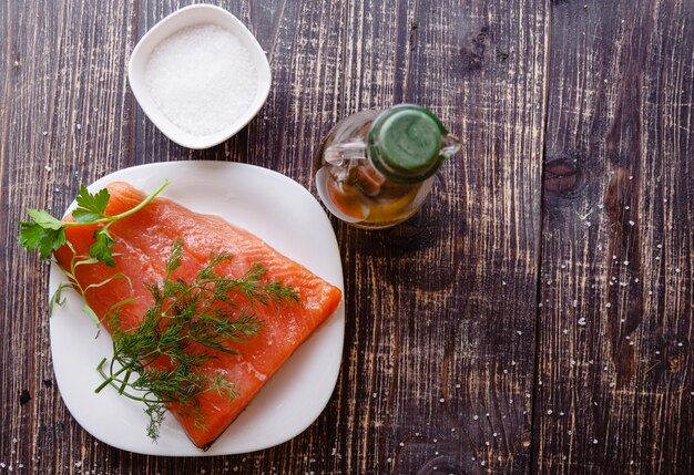 Фото Кусок свежего лосося с веточкой укропа и петрушки на белой тарелке на темном фоне, бутылка оливкового масла и морской соли