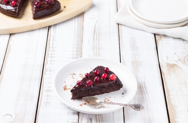 Фото Кусок безглютенового торта в шоколаде, украшенный клюквой, на белой тарелке, на светлом деревянном фоне. здоровые десерты.