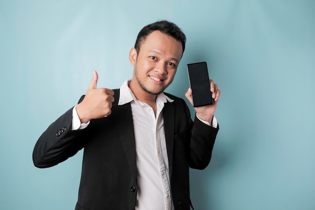 幸せなアジア系のビジネスマンのポートレートが微笑み、青い背景で隔離された黒いスーツを着てit39sの画面にコピースペースを示すスマートフォンを持っている