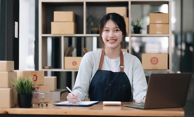 写真 a portrait of a young asian woman ecommerce employee sitting in the office full of packages in the background write note of orders and a calculator for sme business ecommerce and delivery business