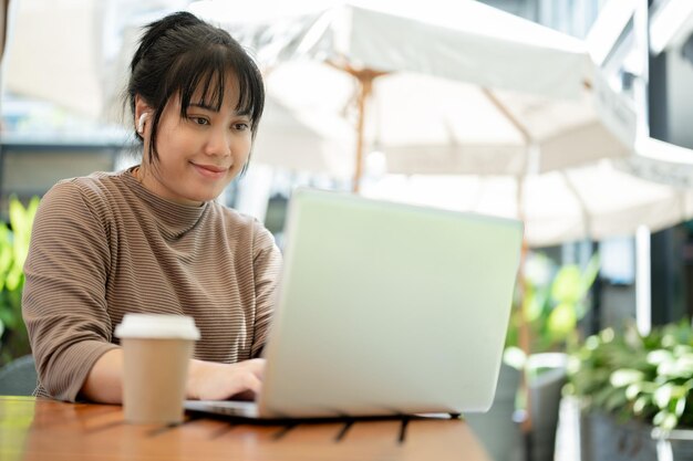 写真 ポジティブなアジア人女性がラップトップを使って都市のカフェでリモートで働いています