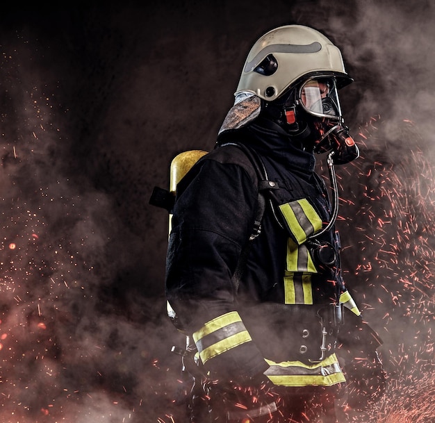 Профессиональный пожарный, одетый в форму и кислородную маску, стоит в огненных искрах и дыме на темном фоне.