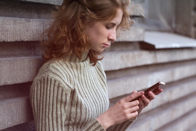 Фото Рыжеволосая девушка с кудрями стоит на улице с телефоном и ищет нужную информацию в интернете