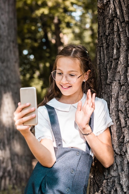 眼鏡をかけた女子高生が、公園の木の近くでスマートフォンでビデオ通話をする 垂直方向のビュー