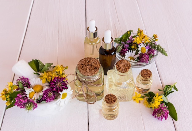 写真 牧草地の薬用植物の選択的な焦点と白い木製のテーブルの上のさまざまなボトルの化粧品の有機およびエッセンシャルオイルのセット