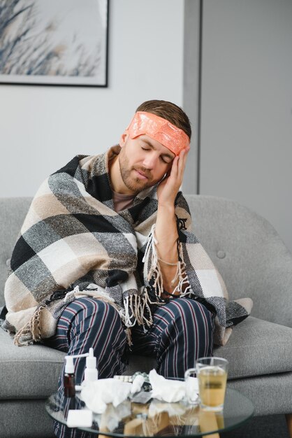 Фото Больной мужчина сидит дома на сером диване с одеялом