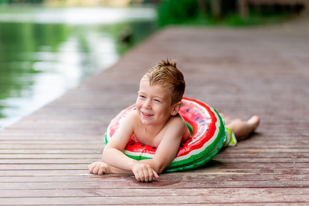 Улыбающийся мальчик с кружком в виде арбуза летом лежит на озере