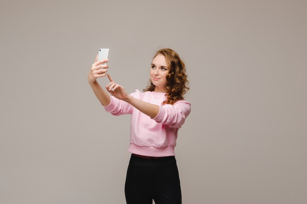 Фото Улыбающаяся счастливая девушка в розовой блузке делает селфи на смартфоне на сером фоне.
