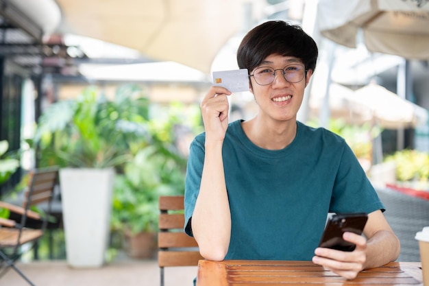Фото Улыбающийся молодой азиатский мужчина показывает свою кредитную карту, сидя за деревянным столом на открытом воздухе