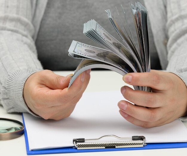 사진 한 여성의 손에 100달러짜리 지폐 더미와 하얀 탁자 위에 있는 나무 돋보기. 예산 분석, 수입 및 지출, 이익 계산