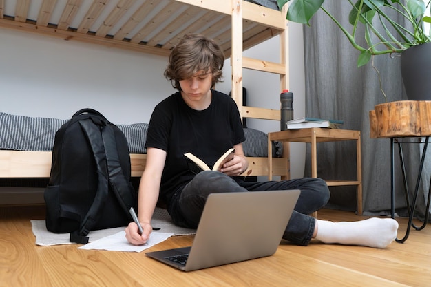 사진 십대 학교 소년 이나 학생 은 노트북 과 열린 책 을 들고 방 에 있는 침대 근처 에 앉아서 숙제를 하고 있으며 종이 한 장 에 펜 을 쓰고 메모 를 하고 있다