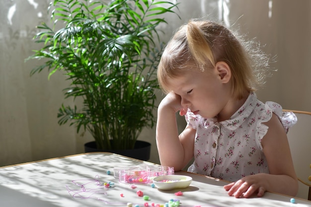 写真 疲れた2歳の女の子がテーブルに座ってビーズビーズを見ています