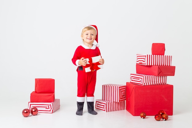Фото Малыш в костюме санты стоит на белом фоне рядом с подарочными коробками на рождество. рождественская концепция, пространство для текста