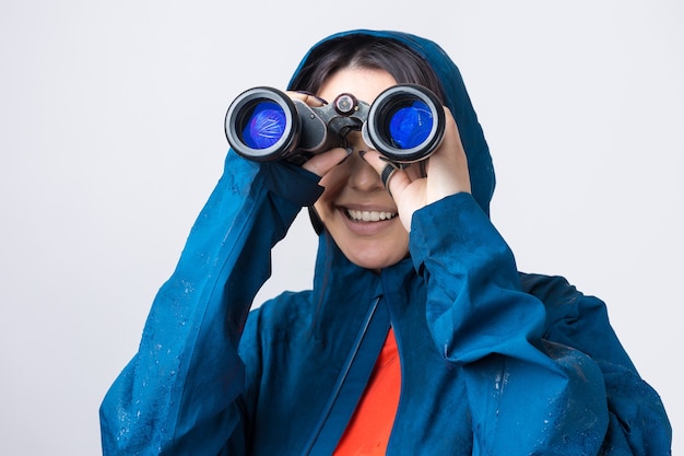 Фото Девушка-туристка в синем плаще держит в руках бинокль и смотрит вдаль, шпионит.