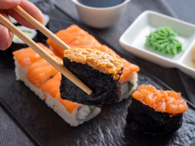 Разнообразные японские роллы и суши на текстурированной черной тарелке. вид сбоку. бамбуковые палочки удерживают один гункан. закрыть вверх