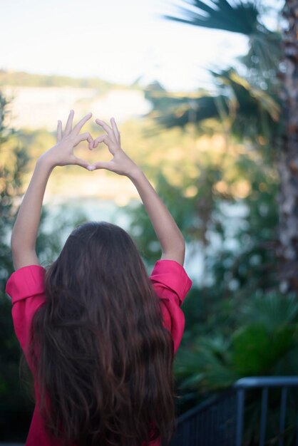 写真 長い髪のピンクのシャツを着た若い女の子のハート型の手の後ろの眺め日没時に子供たちの手で作られたハート熱帯の景色の周りのヤシの木