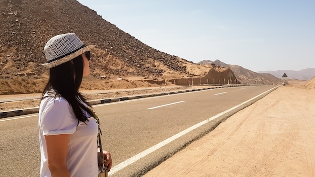 Белая девушка в шляпе стоит у широкой пустынной асфальтовой дороги. задний портрет женщины, смотрящей вдаль на пустой дороге с горами и жарким солнцем. ближний восток. концепция путешествия.